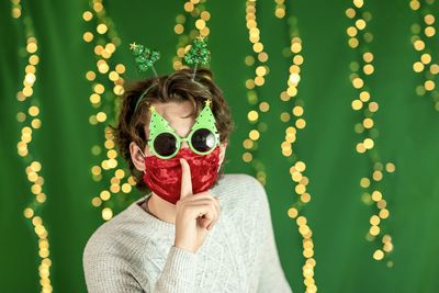 Young man in new year's glasses and red mask on the background of a green wall with new year's light