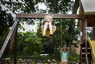 Cute sisters swinging on swings against trees at playground