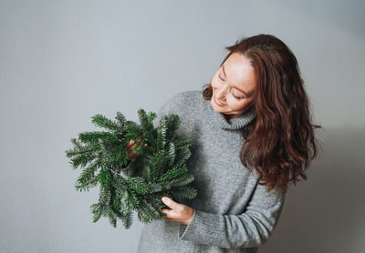 Adult woman in warm grey knitted dress with diy fir christmas wreath in hand on the grey background