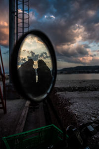 Reflection of man photographing on sea against sky during sunset