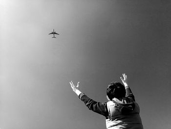 Low angle view of boy looking at airplane flying against sky during sunny day