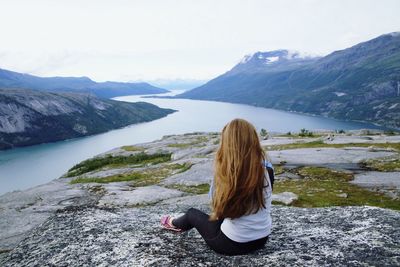 Rear view of woman sitting on rock by river and mountains against sky