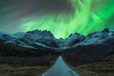 Scenic view of automobile driving on empty mountainous road in winter under night sky glowing with bright green aurora borealis in norway