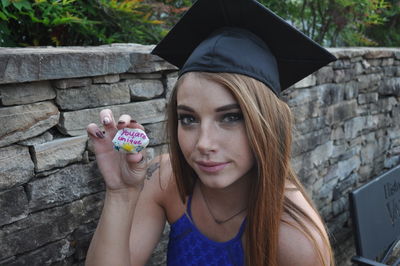 Portrait of girl in mortarboard holding stone with text by wall