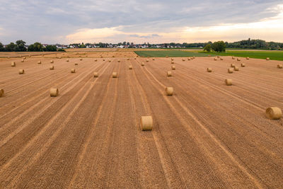 Aerial view of some hay bales on field with some distant houses and trees against sky in back lit