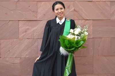 Portrait of young woman in graduation gown holding bouquet against wall