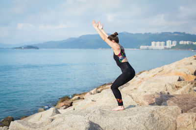 Woman with arms raised exercising while standing on rock by sea