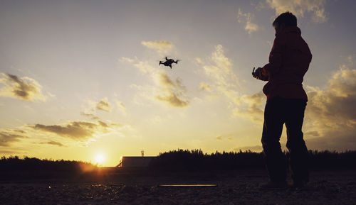 Man flying drone with remote control while standing on land against sky during sunset