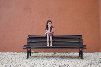 Portrait of girl with hand on chin sitting on bench against wall