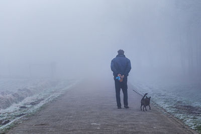 Rear view of man with puppy walking on pathway during foggy weather