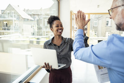 Cheerful businesswoman with laptop doing high-five with male colleague in office