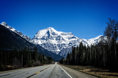 Empty country road amidst trees against snowcapped mountains