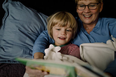 Smiling grandmother reading story book for grandson on bed at home