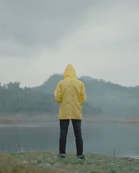 Rear view of man wearing yellow raincoat while standing on riverbank during foggy weather