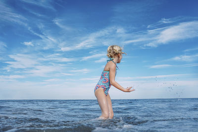 Side view of girl playing while standing in sea against cloudy sky
