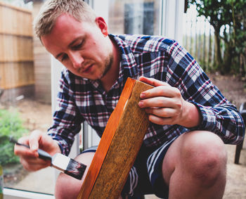 Young man painting wooden plank while crouching in yard