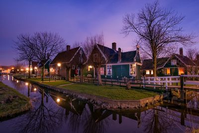 Illuminated houses by canal against sky at dusk