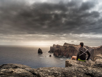 Rear view of man sitting on cliff against cloudy sky