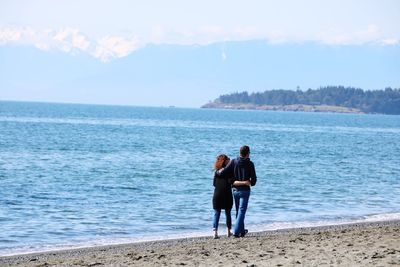 Rear view of couple walking on shore at beach against sky