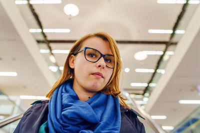 Portrait of young woman wearing eyeglasses while standing against illuminated ceiling