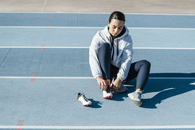 Female athlete in sportswear sitting on racetrack and changing footwear during track and field workout on sunny day on stadium