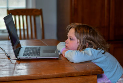 Child in her pajamas relaxes at the table with grandma's laptop, watching kids videos