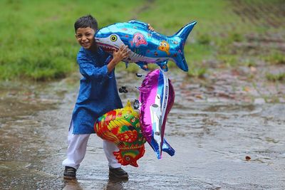 Portrait of smiling boy carrying inflatable animals while standing on footpath during monsoon