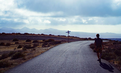 Rear view of man walking on road with roadsign against sky and mountain range