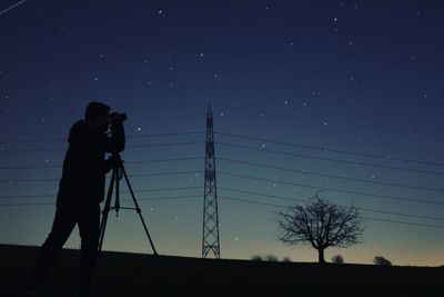 Low angle view of silhouette man photographing electricity pylon against star field at night
