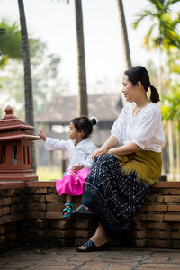 Full length of mother and daughter sitting on retaining wall against building