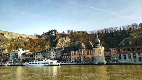 Buildings by meuse river against sky in dinant city