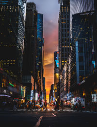 Illuminated city street by buildings against sky during sunset