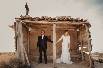 Full body man and woman in elegant clothes holding hands while standing in weathered wooden shelter on wedding day in bardenas reales natural park in navarra, spain