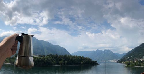 Cropped hand of person holding pitcher by lake against cloudy sky