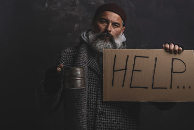 A poor homeless man with placard and a pot for alms on a dark background person