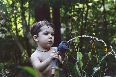 Shirtless boy holding hose by plants on field