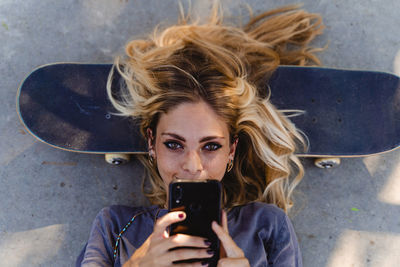 Blond woman using smart phone while lying on skateboard over road
