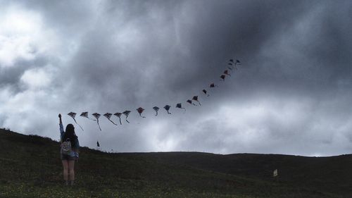 Rear view of woman flying kites on mountain against cloudy sky