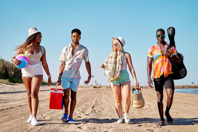 Full body multiracial men and women in sunglasses and hat strolling on sand against cloudless blue sky while spending time on beach on summer day