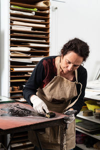 Mature female artist in apron and gloves covering engraved plate with ink while creating artwork in professional workroom