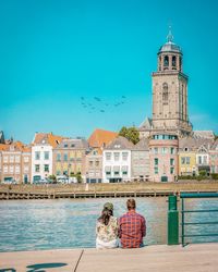 Rear view of couple looking at buildings while sitting by river against clear blue sky in city