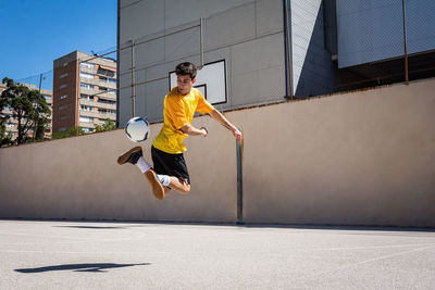 Young male player kicking football while practicing soccer against building