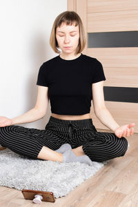 Full length of young woman meditating while sitting on wooden floor