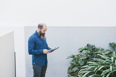 Busy male worker standing in creative workspace with plants and browsing tablet while working on new project