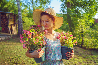 Smiling woman carrying plants while standing in yard against trees