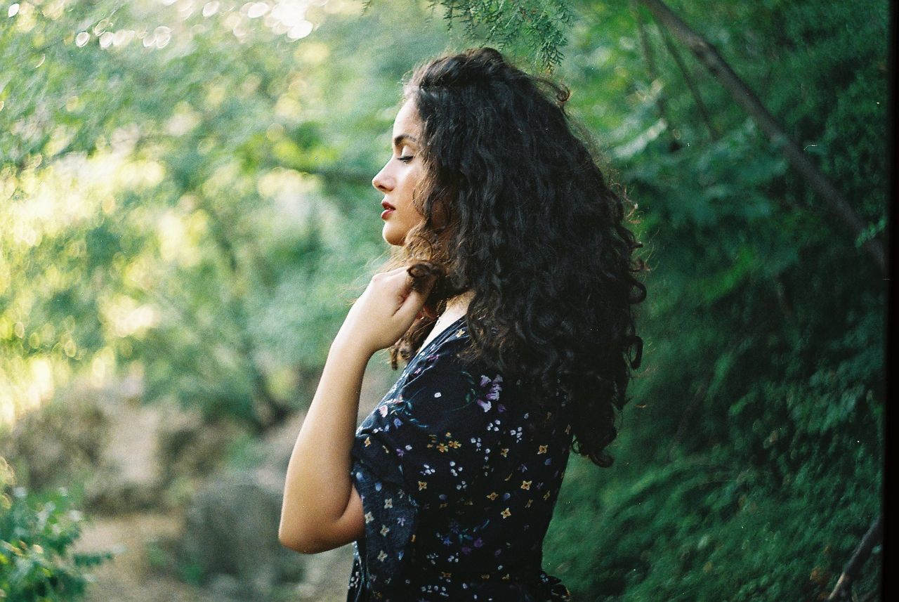 one person, hairstyle, women, adult, tree, long hair, nature, young adult, forest, plant, curly hair, sunlight, green, land, photo shoot, portrait photography, portrait, standing, contemplation, outdoors, looking, lifestyles, dress, person, fashion, side view, brown hair, waist up, smiling, leisure activity, focus on foreground, beauty in nature, day, emotion, environment, happiness, looking away, tranquility, casual clothing, black hair, summer, clothing, female