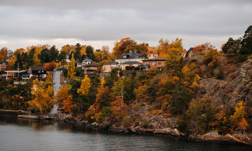 Trees and houses by river against sky during autumn