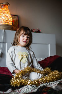 Upset girl in beige pajamas holds gold christmas decoration sitting on the bed