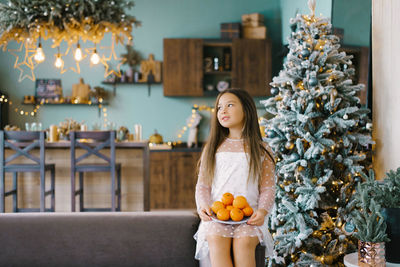 A cute happy little girl in a beautiful white dress is sitting on the sofa next to a christmas tree