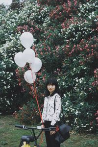Portrait of young woman with balloons and bicycle standing against flowering plants in park